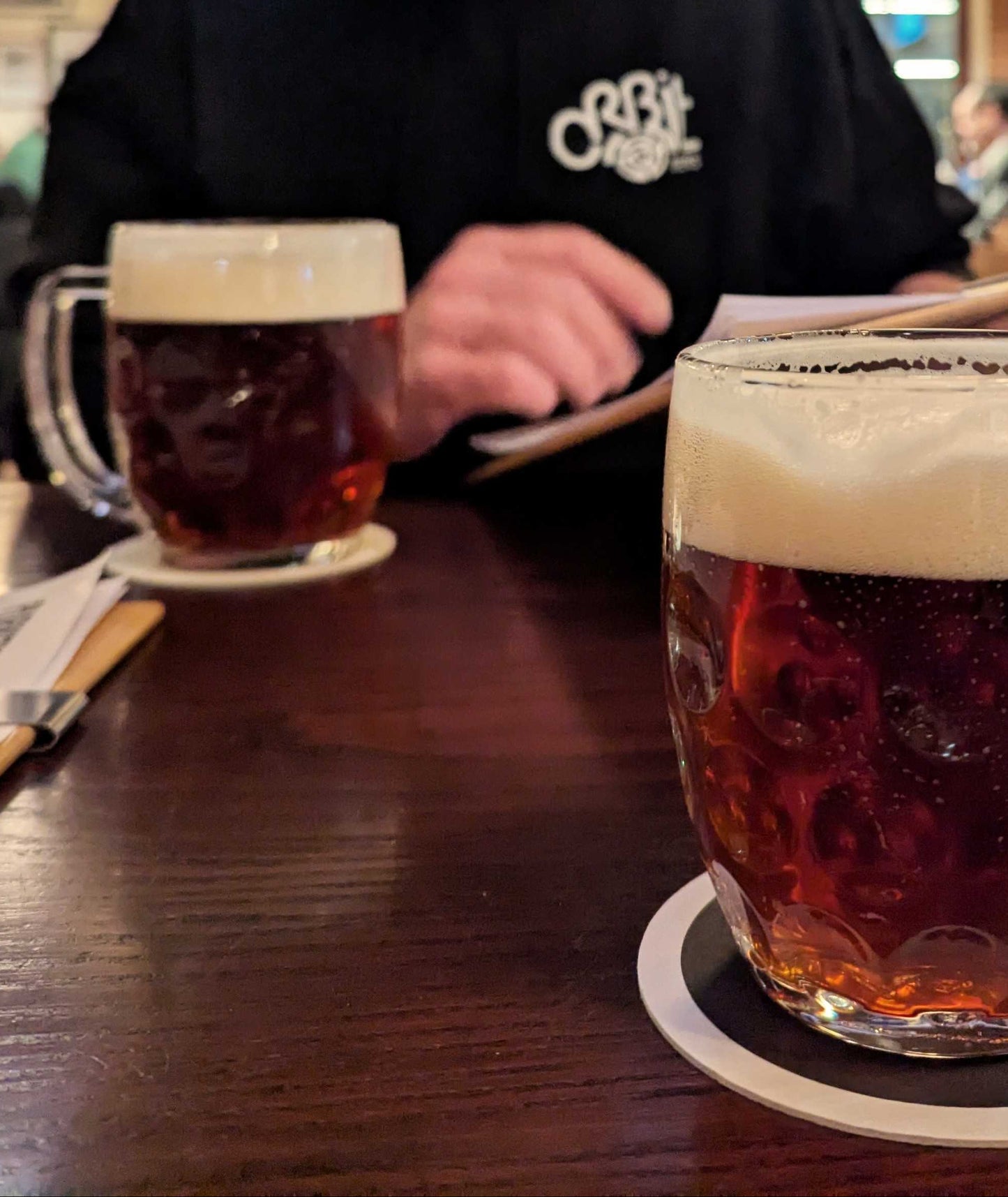 Two mugs of beer on a bar counter with a person in the background.