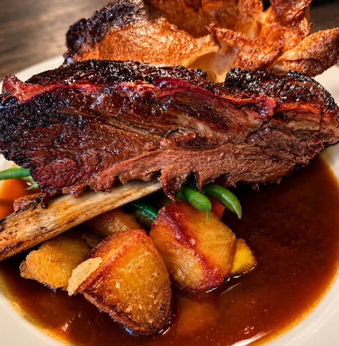 A close-up of a plated cooked meat dish from Dragon Flame BBQ with a large piece of roasted beef or lamb on a bone, served over potatoes and green beans in a red wine gravy.